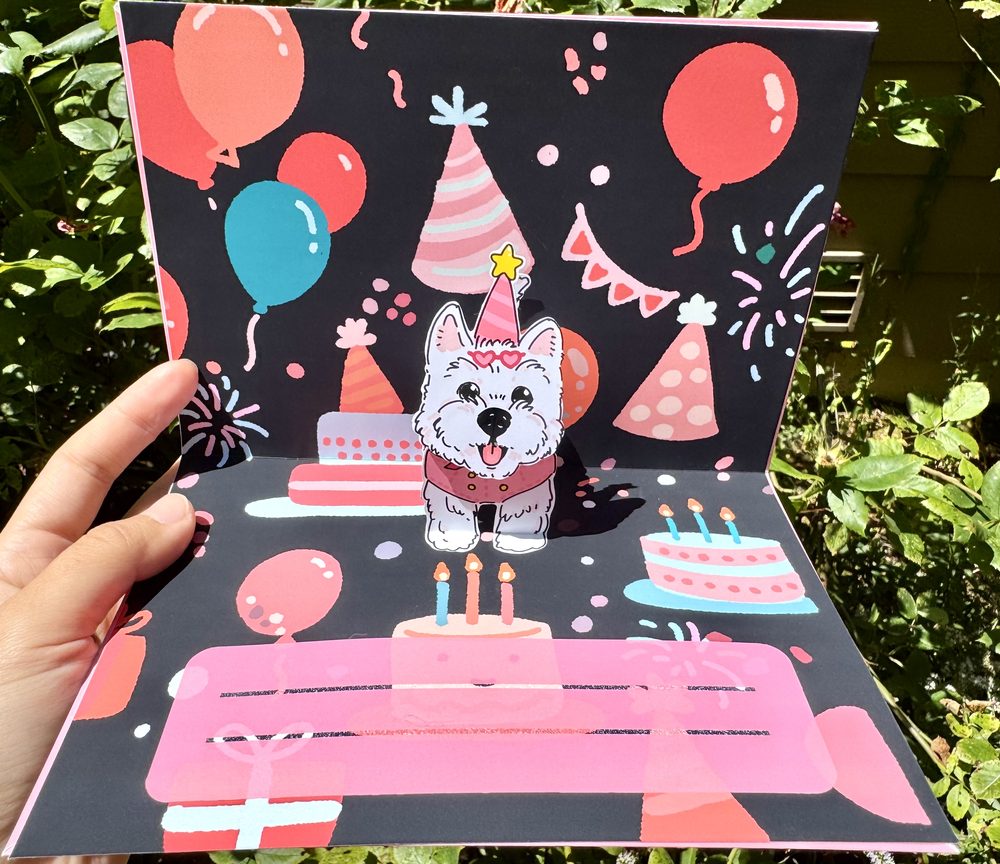 Pop-up birthday card opened outdoors: a white westie in a party hat stands up from the card in front of balloons and cake